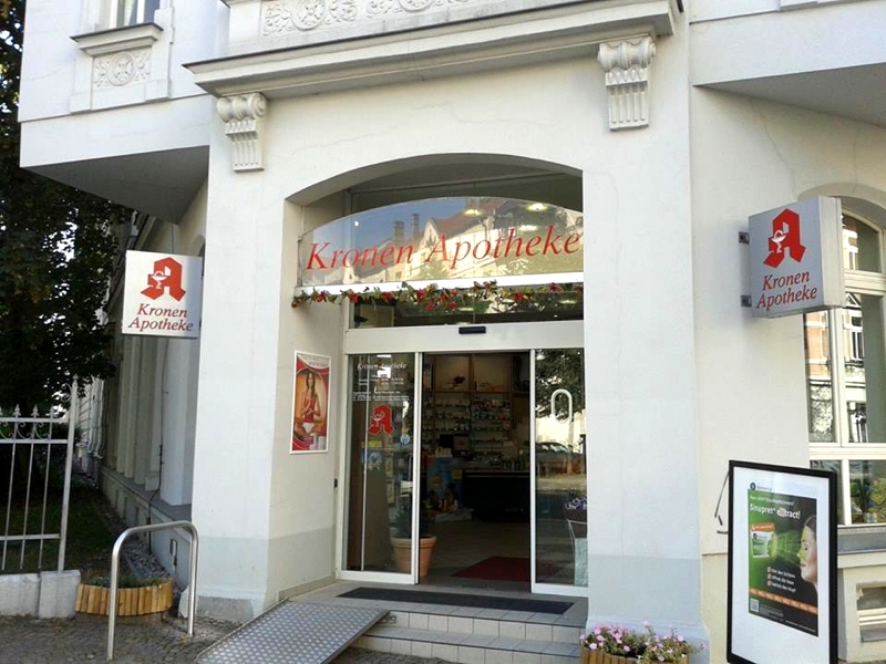 Außenansicht der Kronen Apotheke in Leipzig. Das Bild zeigt den Eingangsbereich der Apotheke mit automatischer Schiebetür, Apothekenschildern und barrierearmem Zugang über eine Rampe. Durch den Eingang ist der Verkaufsraum teilweise sichtbar.